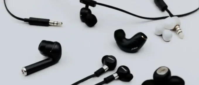 A high-angle, detailed shot of various earbuds, including a pair of classic wired earbuds, a set of wireless earbuds with a connecting neckband, and a sleek, modern true wireless earbud with its charging case, arranged on a neutral background to illustrate the different types of headphone technology.